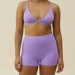 Woman wearing a purple triangle swim top and swim shorts set on a beige background